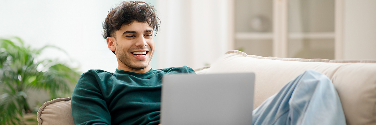 A photographic style image of a male in his early twenties using a computer while reclining on a couch.