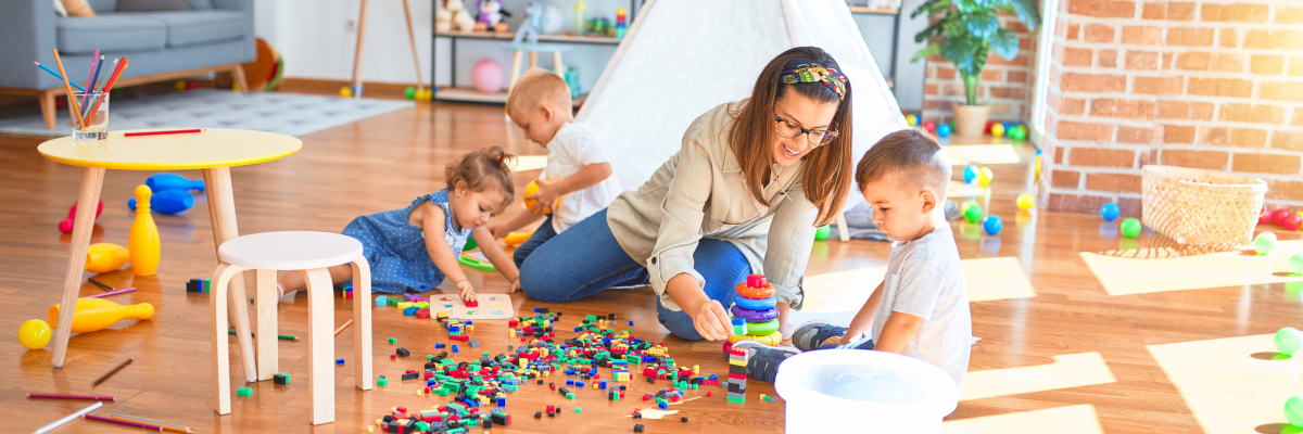 A photographic image of a teacher and toddlers playing with toys in a home