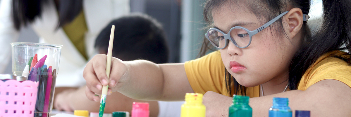 Photographic style image of a young girl with Down's syndrome painting a picture at school